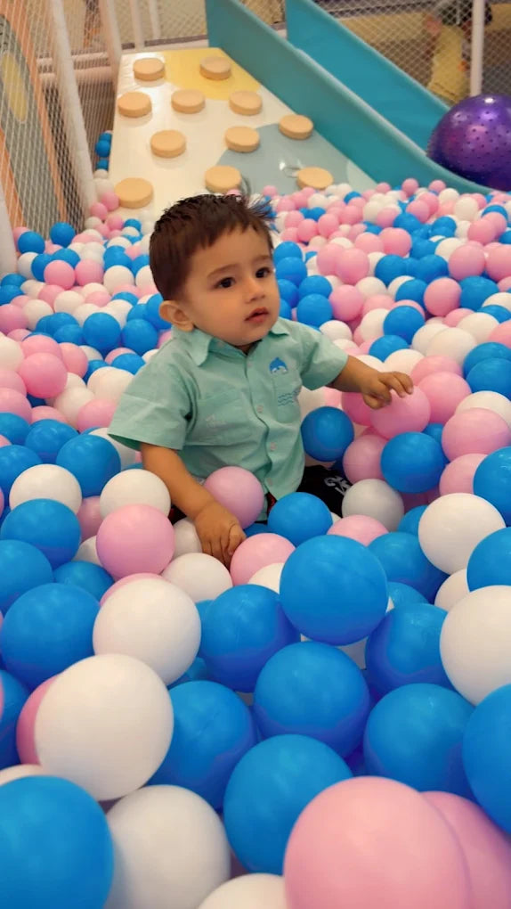 A young child sits in a ball pit filled with blue, pink, and white balls.
