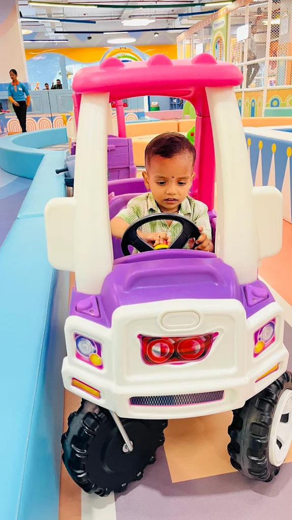 A young child sits in a toy car at an indoor playground. The child is holding the steering wheel.