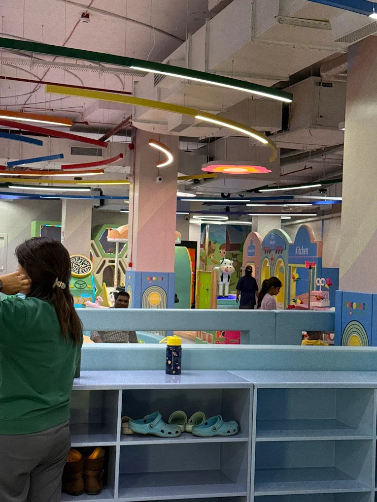 A woman stands in a colorful indoor play area, looking towards children playing. Shelves with shoes are in the foreground.
