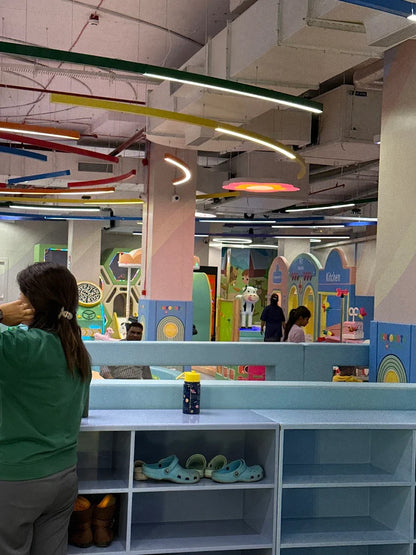 A woman stands in a colorful indoor play area, looking towards children playing. Shelves with shoes are in the foreground.