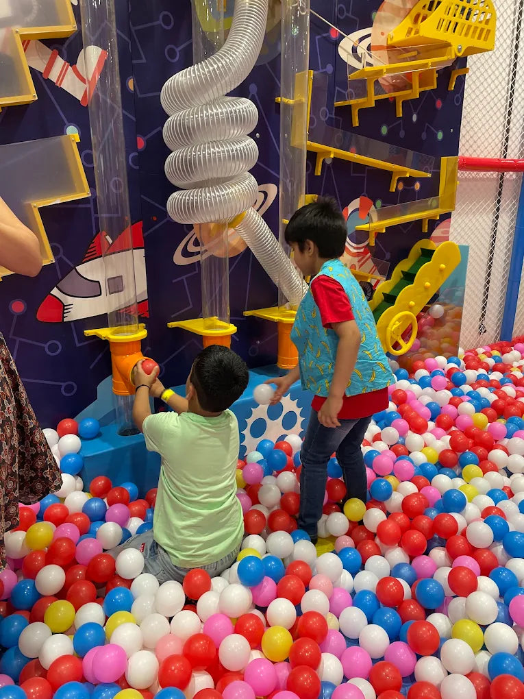 Two young boys play in a ball pit with a colorful, interactive wall featuring tubes and slides.