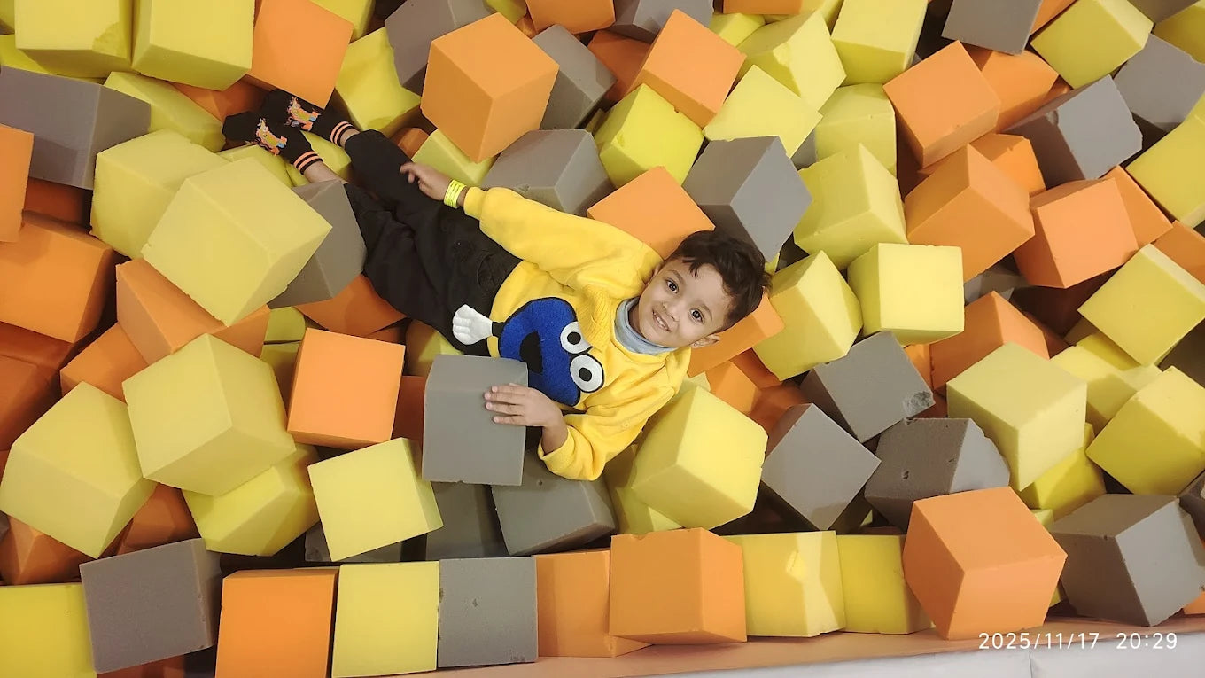 A young boy smiles while lying in a ball pit filled with colorful foam cubes.