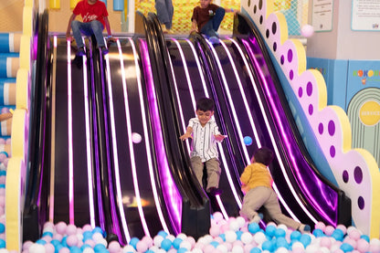 Children slide down a colorful, illuminated playground slide into a ball pit.