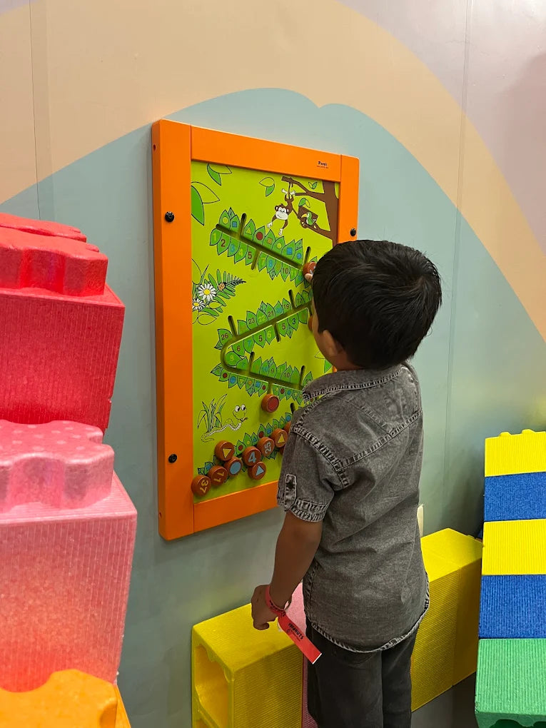 A young boy plays a monkey-themed bead maze game on a wall in a colorful playroom.