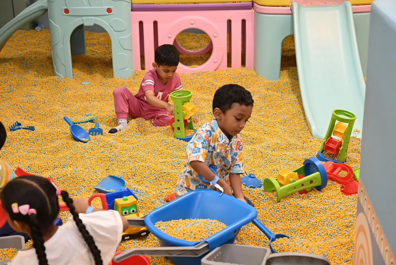 Children play in a ball pit with toys, including a blue wheelbarrow and sand molds.