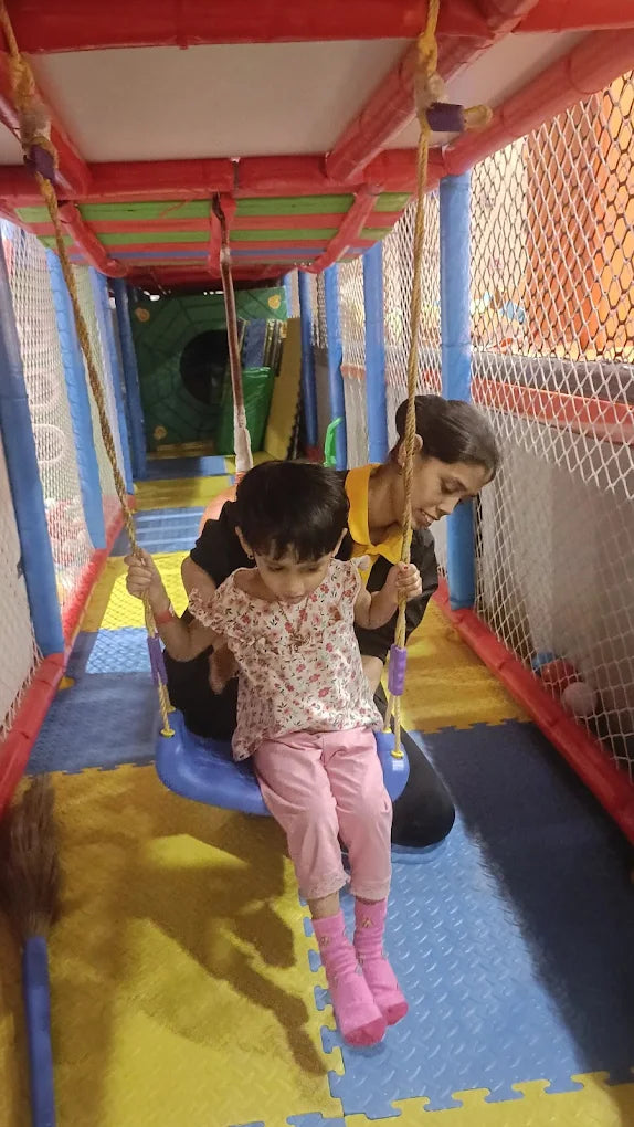 A young child swings on a blue swing while an adult supervises in a colorful indoor playground.