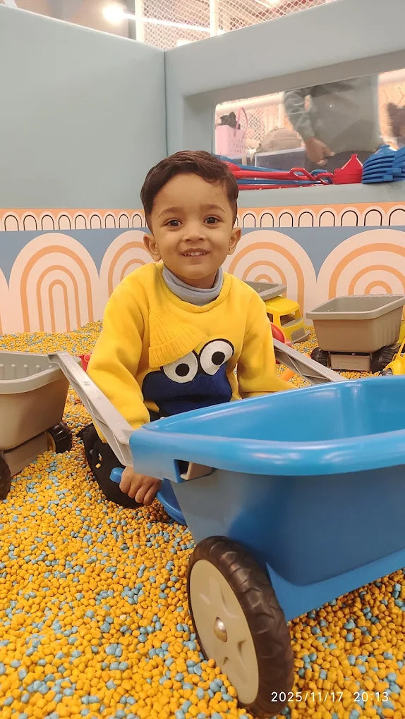 A young boy in a yellow sweater sits in a ball pit with a blue toy wheelbarrow.