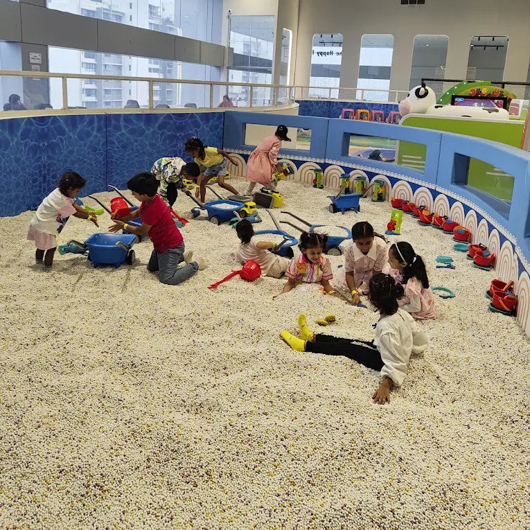 A group of children playing in a large indoor ball pit play area with blue walls, toy vehicles, and outdoor views in the background.