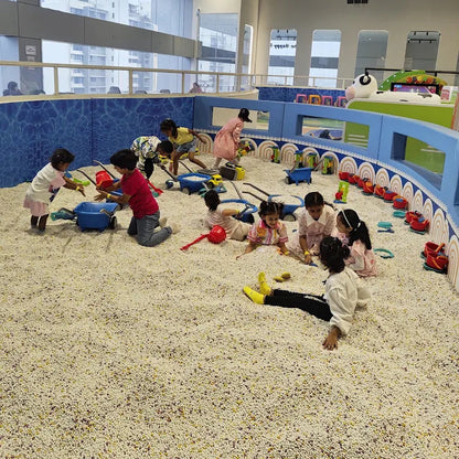A group of children playing in a large indoor ball pit play area with blue walls, toy vehicles, and outdoor views in the background.