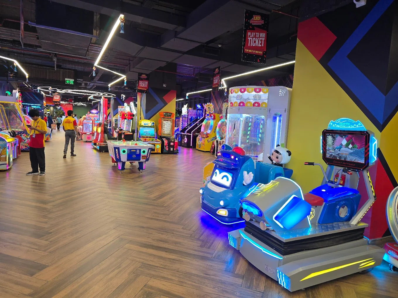 An arcade with various game machines and a few adults playing.
