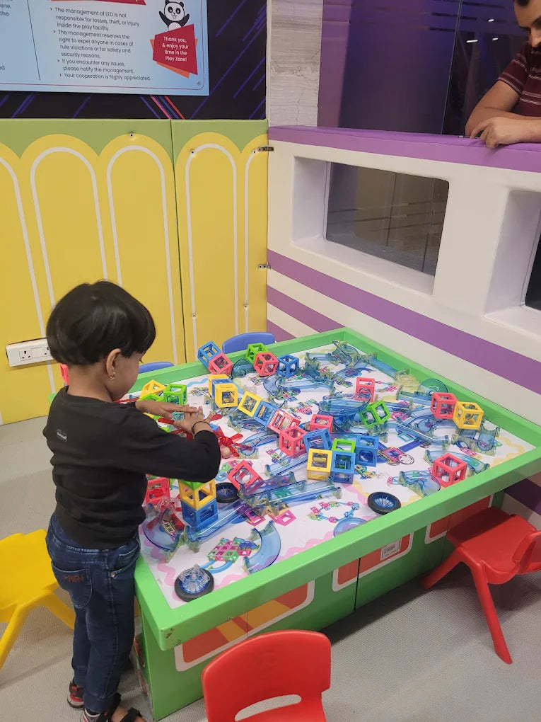 A young child plays with colorful building blocks on a large table in a play area.