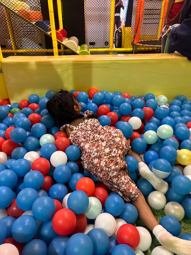 A young child lies in a ball pit, surrounded by blue, red, and white balls.