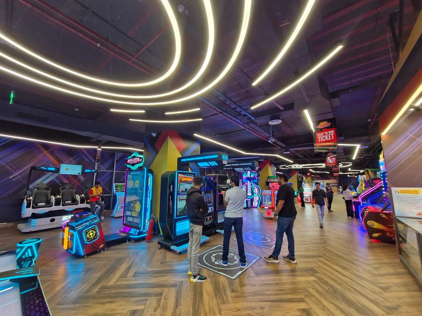 People play arcade games in a brightly lit entertainment center with neon lights and colorful machines.