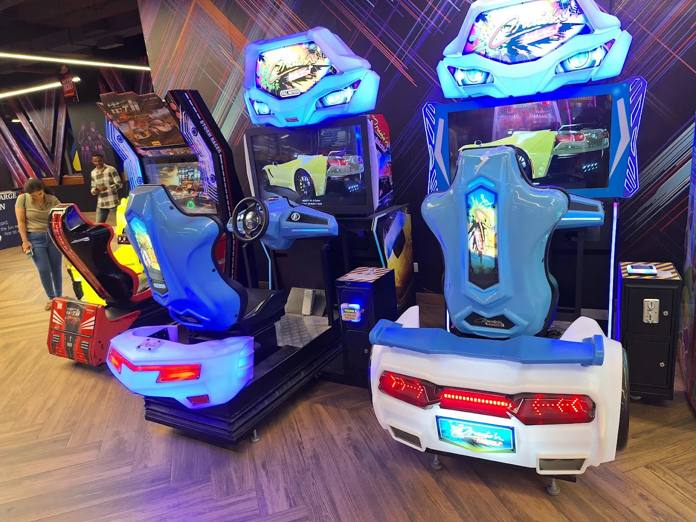 Arcade racing game machines with colorful lights and screens, with young adults playing.