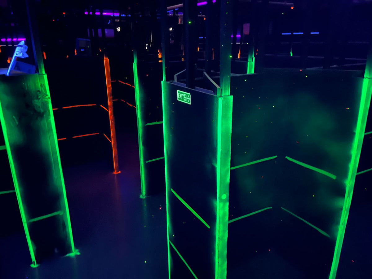 A dark laser tag arena with neon green and orange lights, featuring maze-like walls and an exit sign.