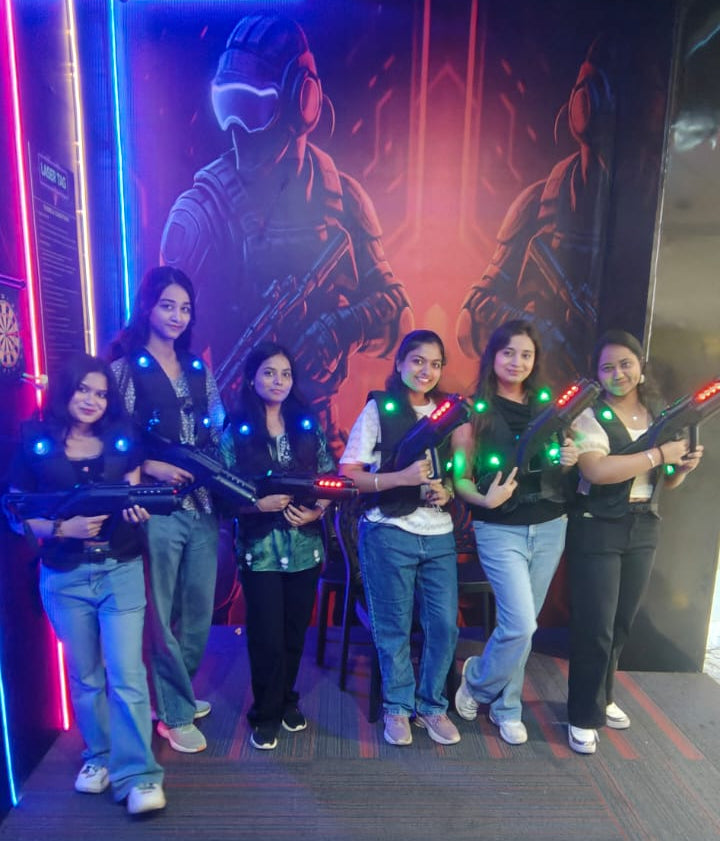 Young women in laser tag vests and holding phasers stand in a dimly lit arena with a futuristic mural.