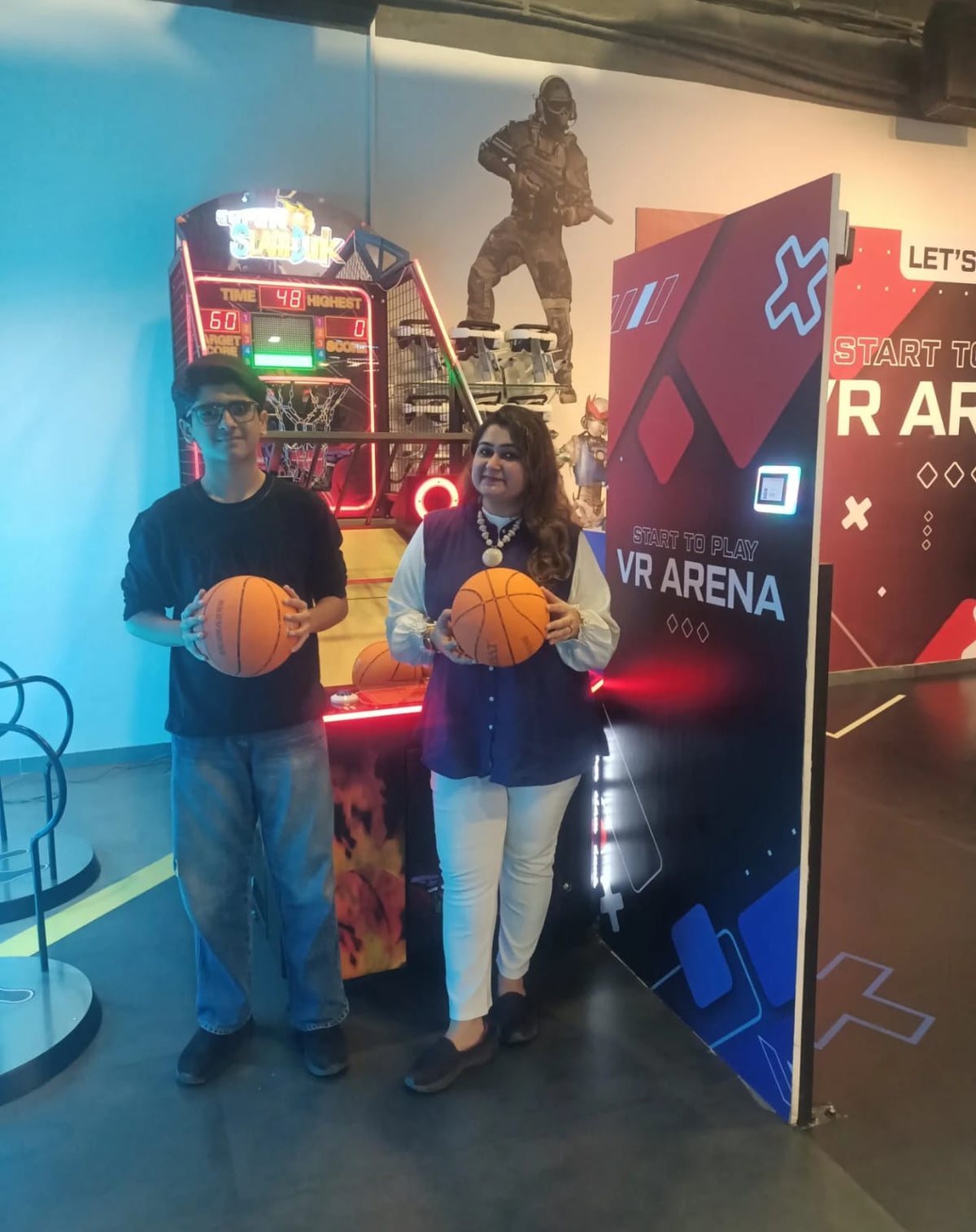 Two people hold basketballs in front of a VR arena and a basketball arcade game.