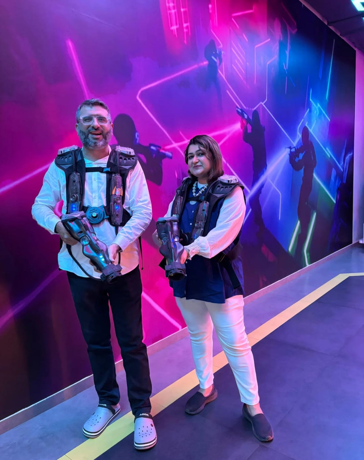 A man and woman in laser tag vests and gear stand in front of a neon-lit wall.