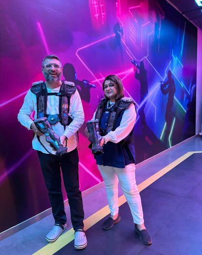 A man and woman in laser tag vests and gear stand in front of a neon-lit wall.
