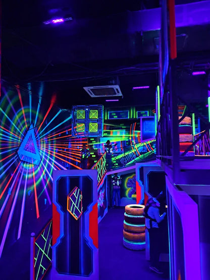 A group of young people play laser tag in a neon-lit arena with glowing walls and obstacles.