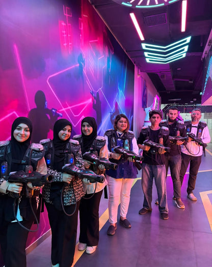 A group of adults in a laser tag arena, holding phasers and wearing vests, with neon lights in the background.