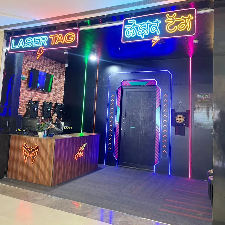 A young woman works at the counter of a brightly lit laser tag arena with neon signs.