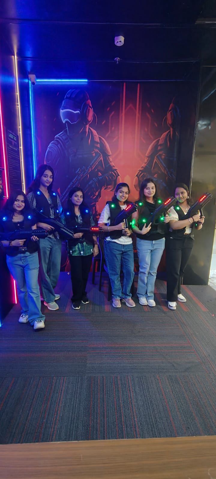 Young women in laser tag vests and holding phasers pose in a dimly lit arena with a futuristic mural.