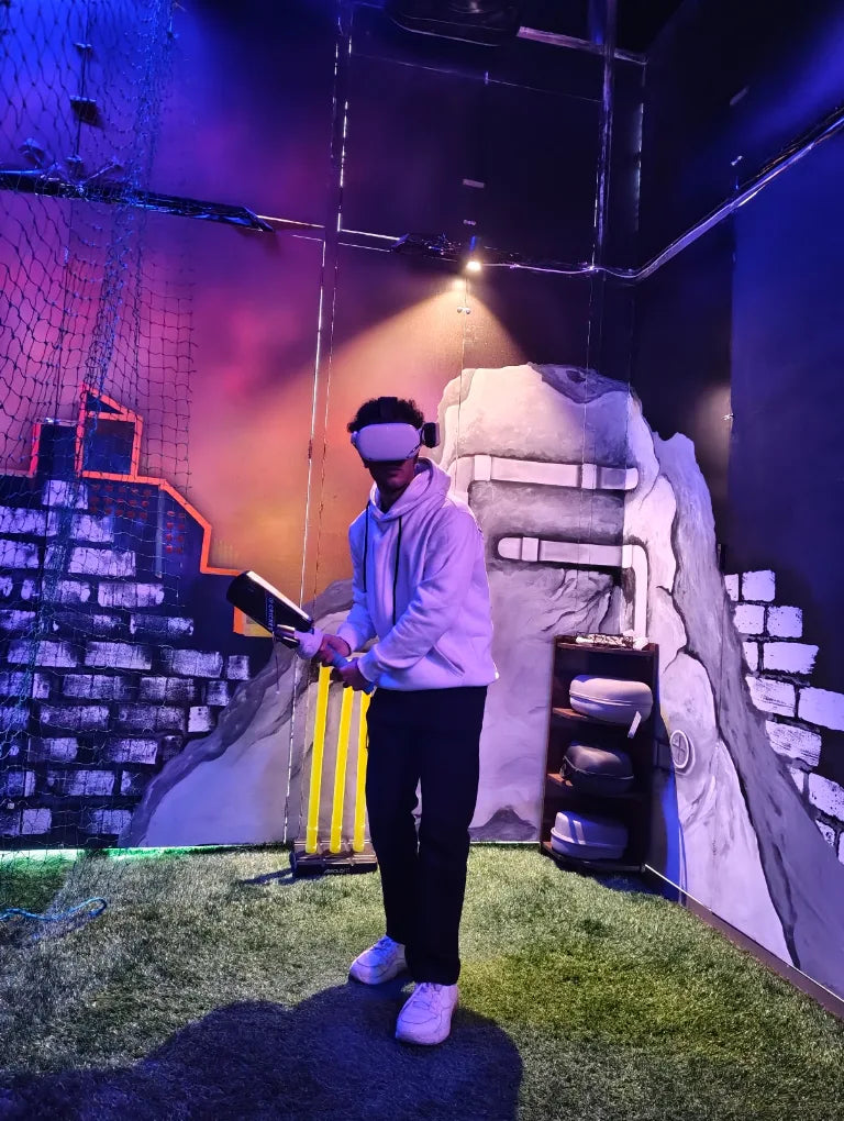A young person in VR goggles plays a virtual cricket game in a dimly lit, colorful room.
