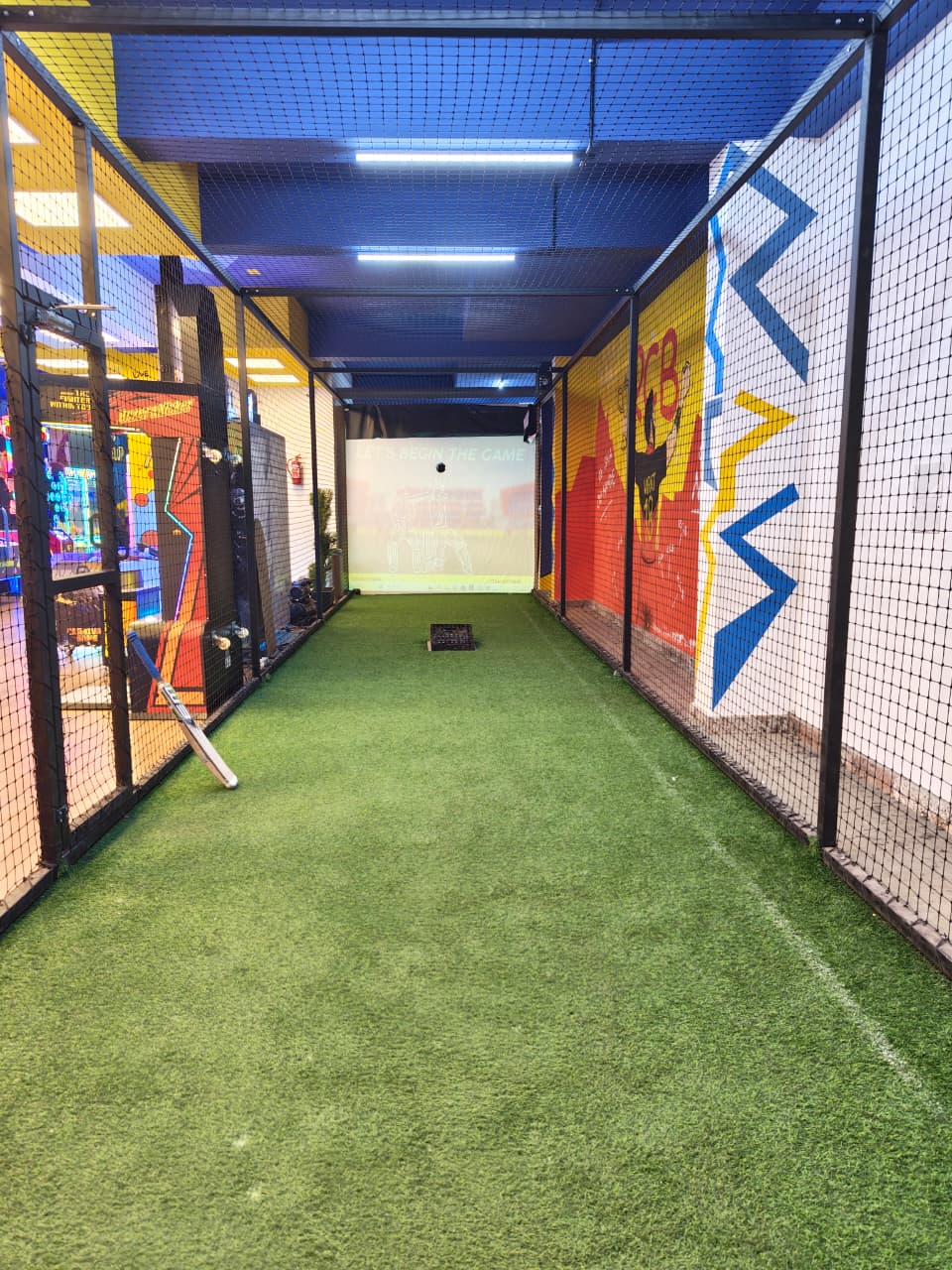 Indoor batting cage with artificial turf, a screen displaying "LET'S BEGIN THE GAME," and a cricket bat.