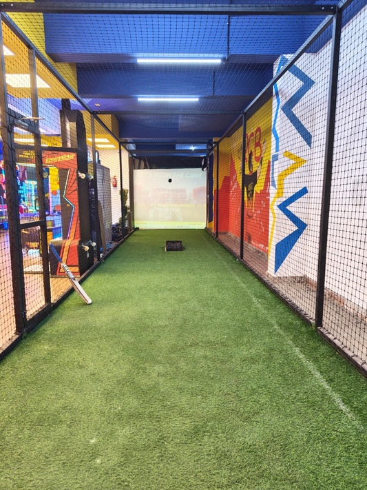 Indoor batting cage with artificial turf, a screen displaying "LET'S BEGIN THE GAME," and a cricket bat.