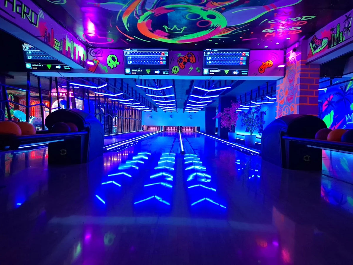 A neon-lit bowling alley with glowing blue lanes and vibrant graffiti art on the ceiling.