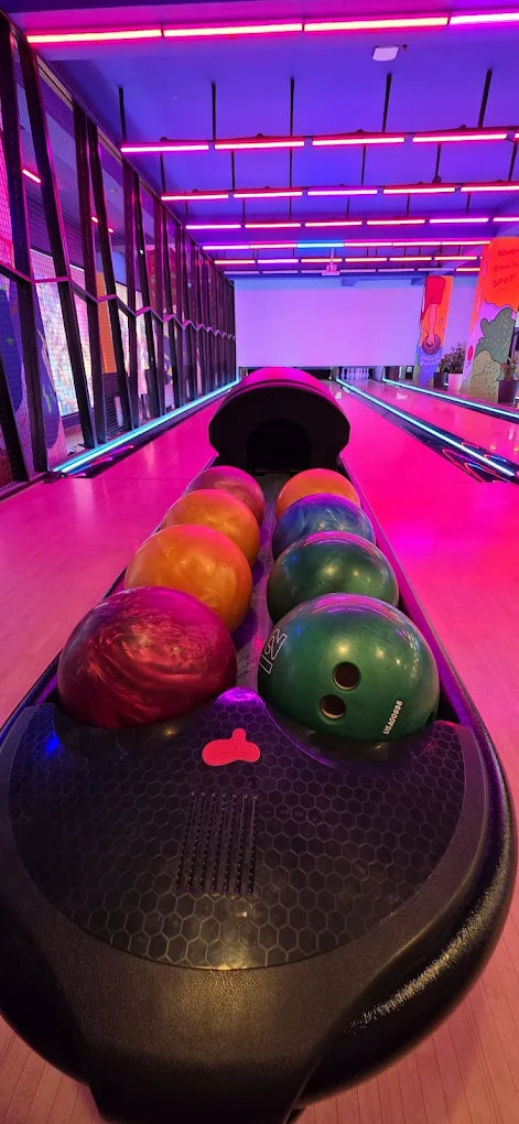 Bowling balls lined up on a lane in a bowling alley with neon pink lighting.