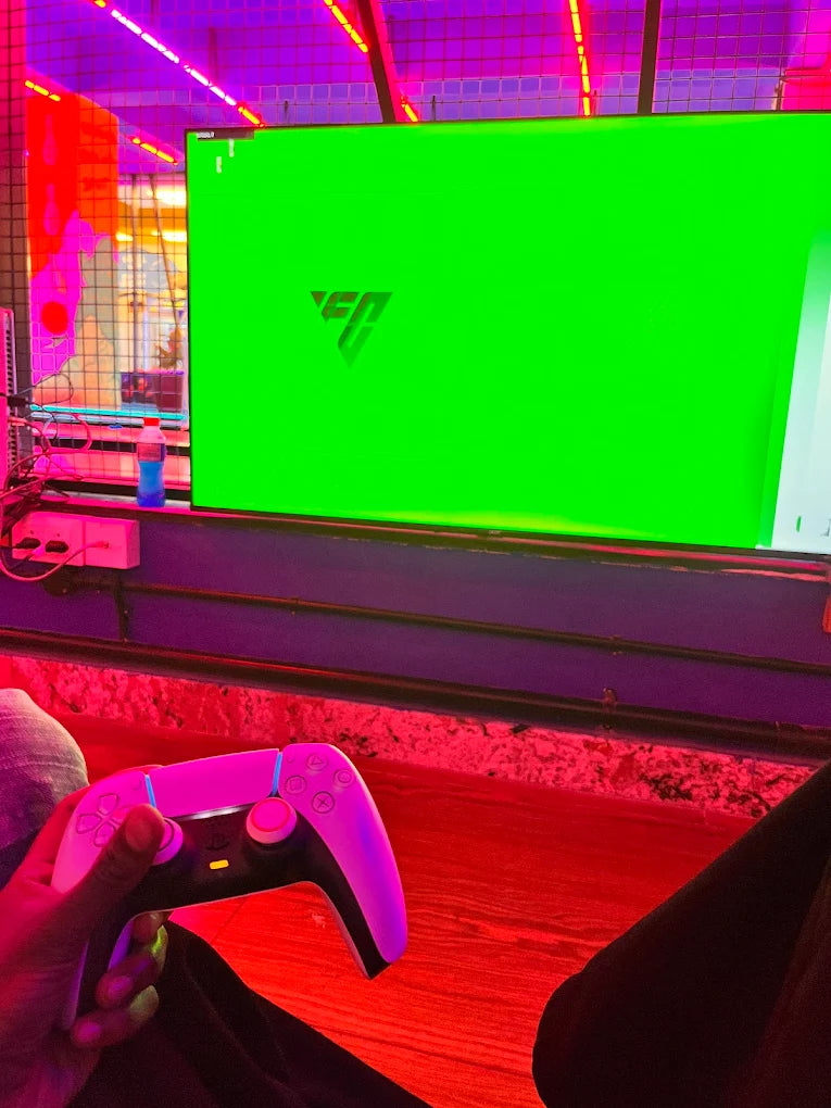 A person holds a white game controller in front of a large green screen displaying a logo. The scene is lit with vibrant pink and red lights.