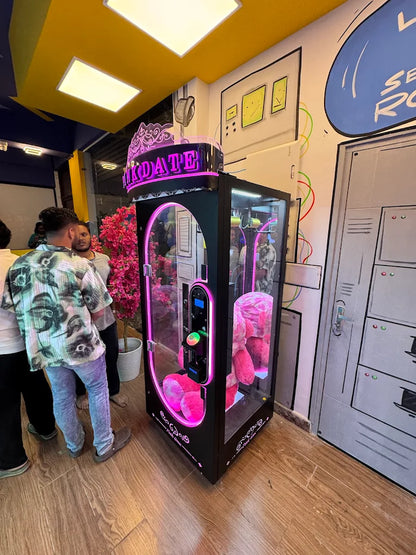 A claw machine filled with pink stuffed animals is in a brightly lit arcade with young adults.