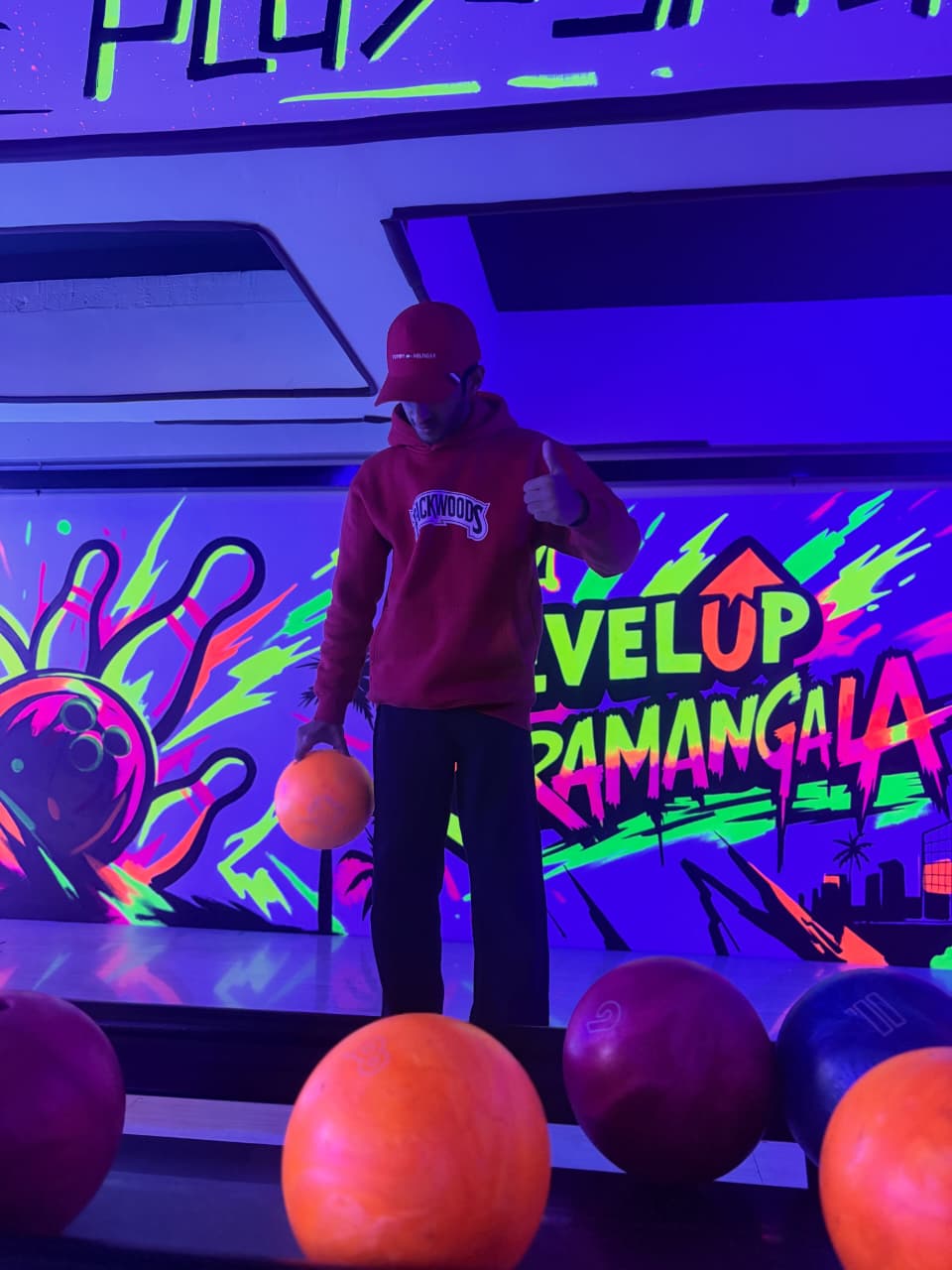 A young person in a red hoodie and hat holds a bowling ball in a neon-lit bowling alley.