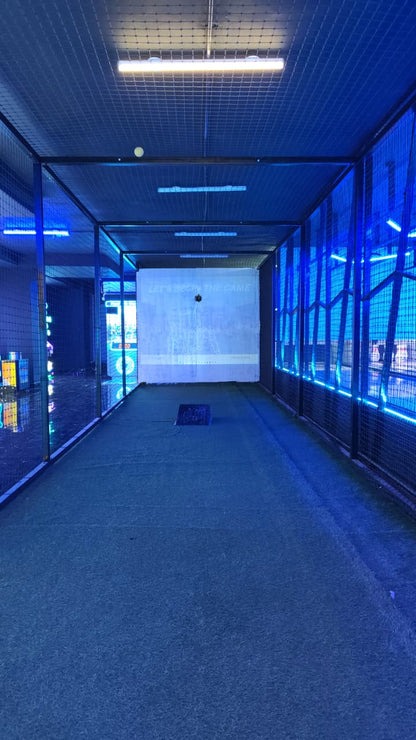 A batting cage with a projected screen and blue lighting. A baseball is visible in the air.