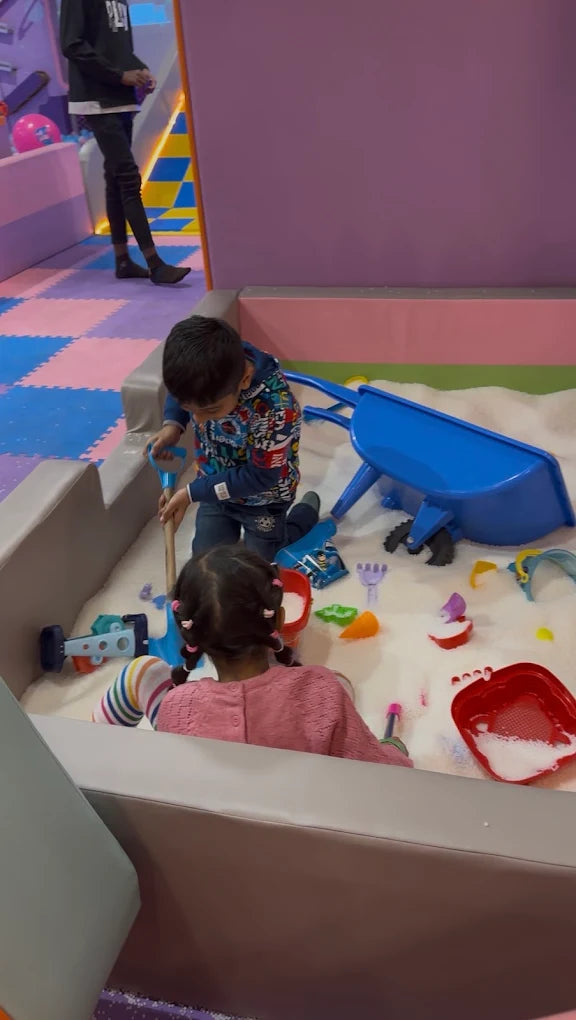 Children play in a sandpit with toys, including a blue wheelbarrow and shovels.