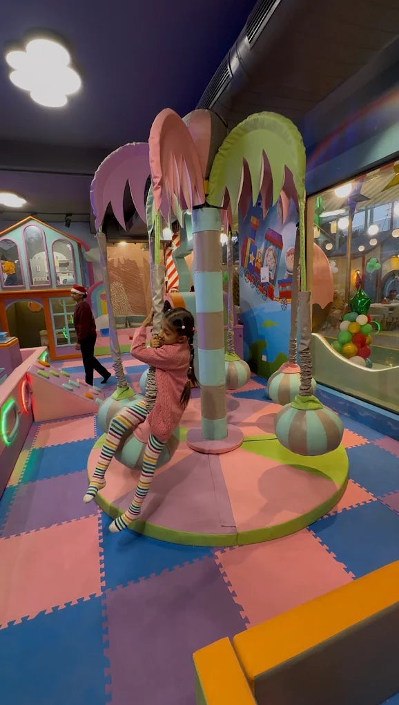 A young girl swings on a rope in a colorful indoor playground. The play area features soft mats, palm tree-like structures, and a playhouse.