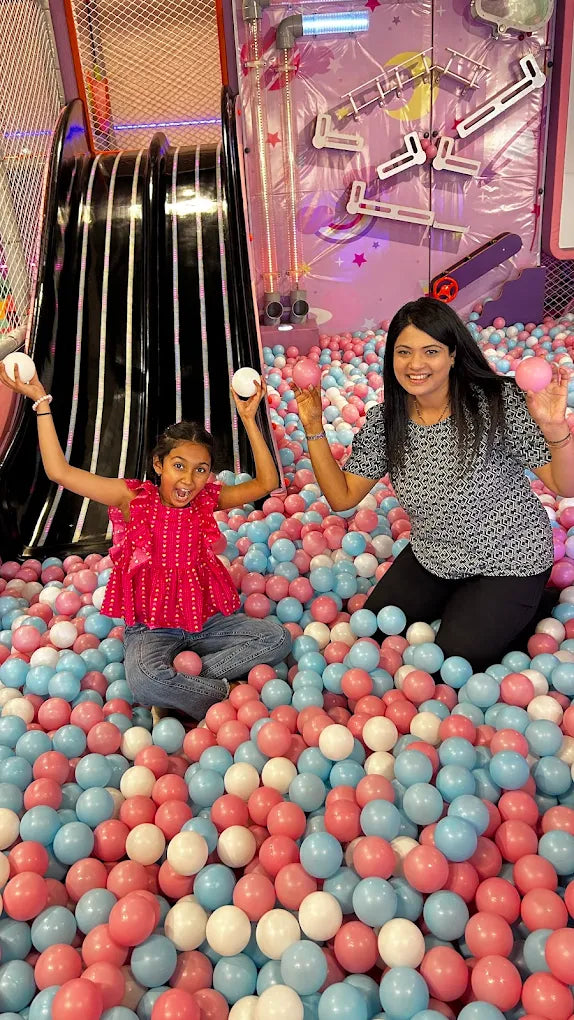 A young girl and woman play in a ball pit with pink, blue, and white balls.