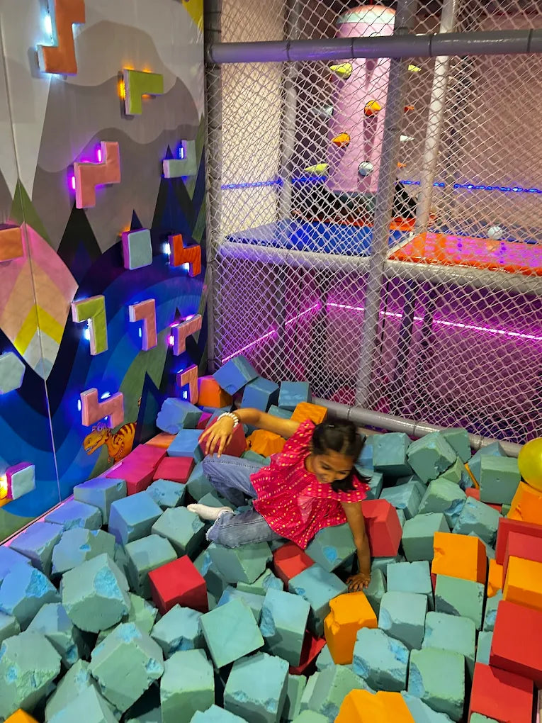 A young girl plays in a ball pit filled with large foam blocks. The colorful pit is part of an indoor play area with climbing walls.