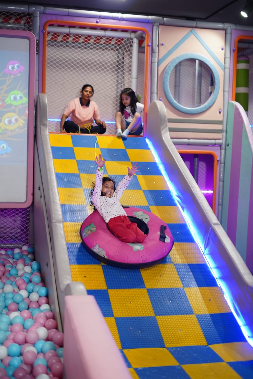 A young girl slides down a colorful playground slide in a tube, while two women watch from above.