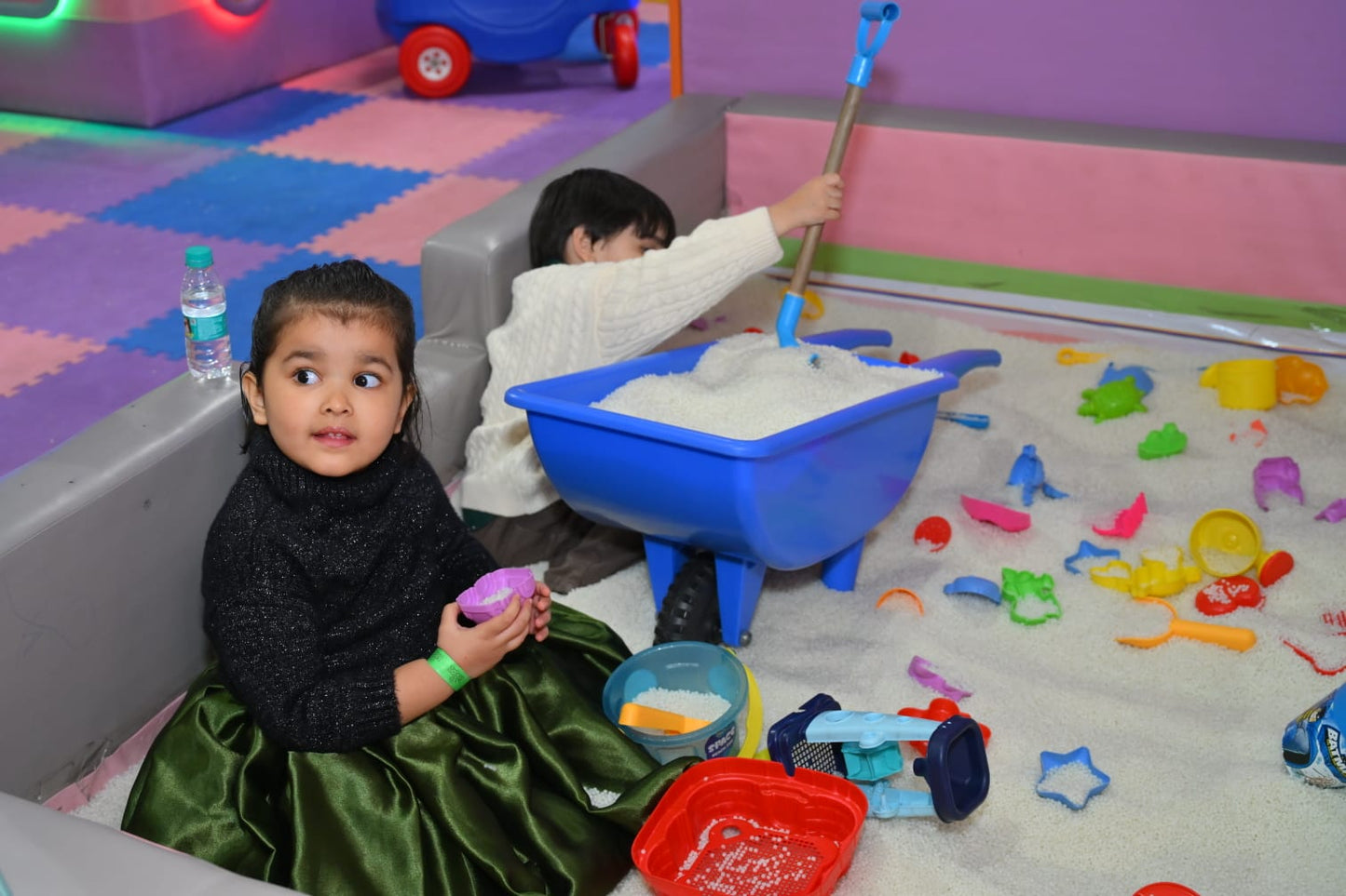 Two young children play in a sandbox filled with white beads and toys.