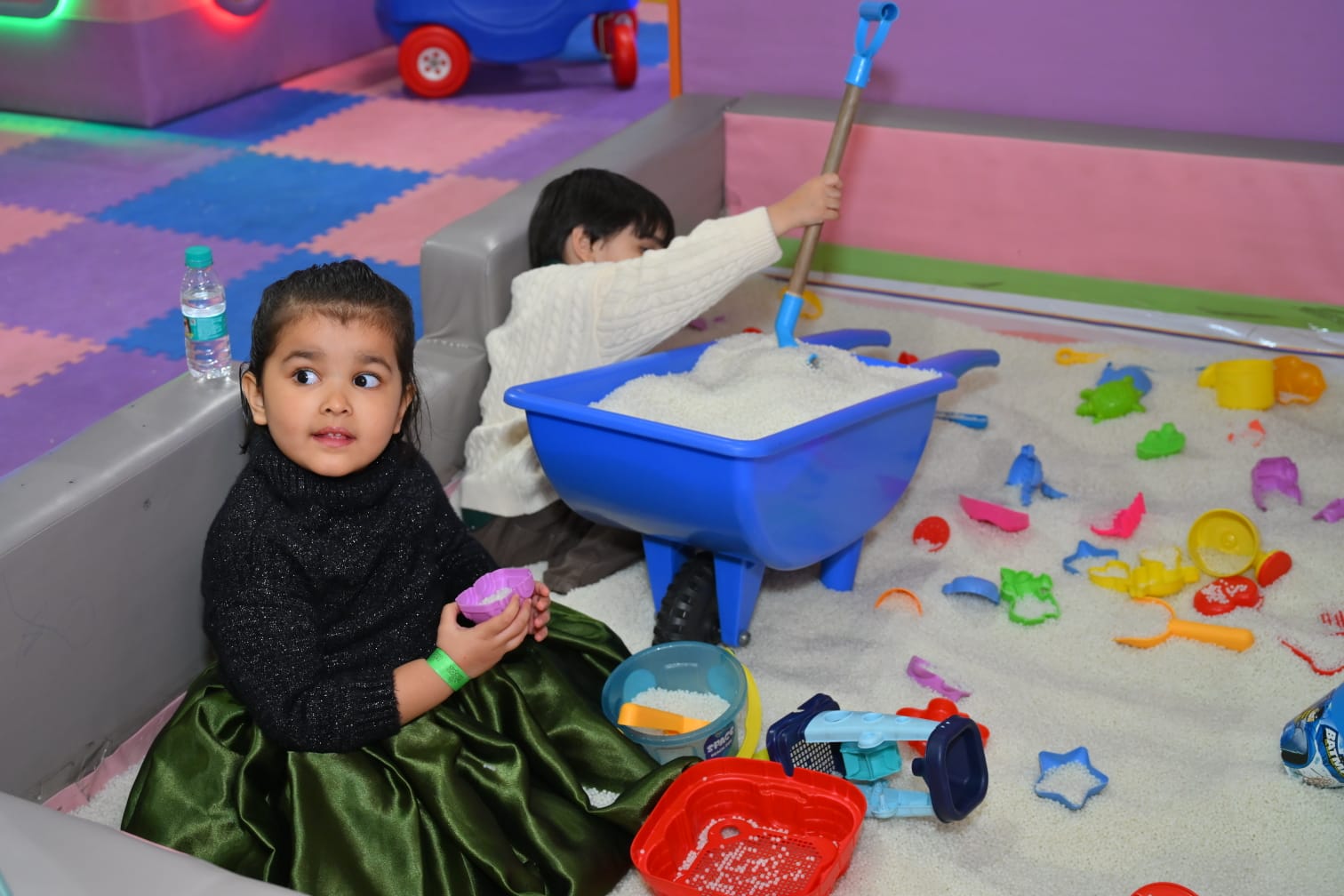 Two young children play in a sandbox filled with white beads and toys.
