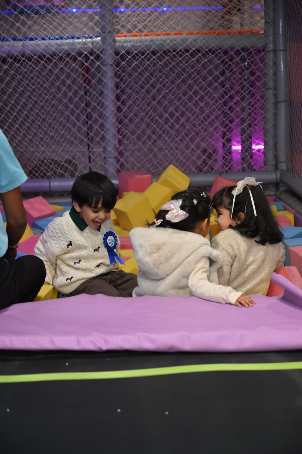 Three young children sit in a ball pit, two facing each other and one looking down and laughing.