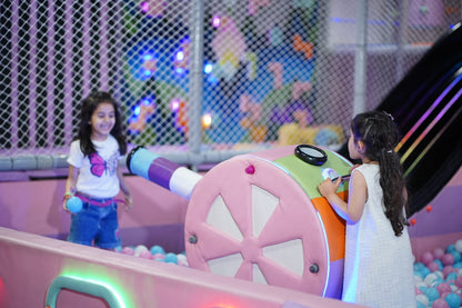 Two young girls play in a colorful indoor playground with a ball pit and a toy cannon.