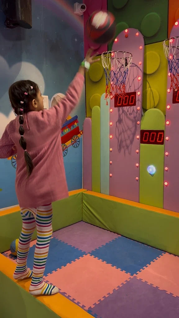 A young girl in colorful striped tights plays basketball at an arcade. She is reaching up to shoot a ball towards a hoop.