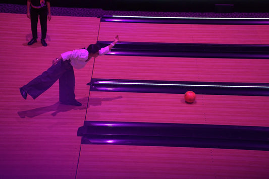 A young person bowls a ball down a lane, with another person standing in the background.
