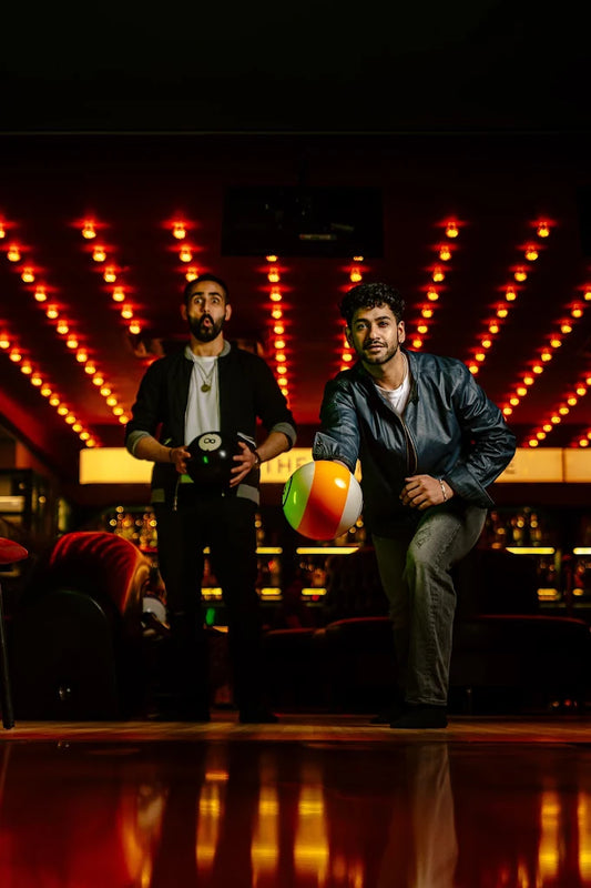 Two young men play bowling in a dimly lit alley with red lights.