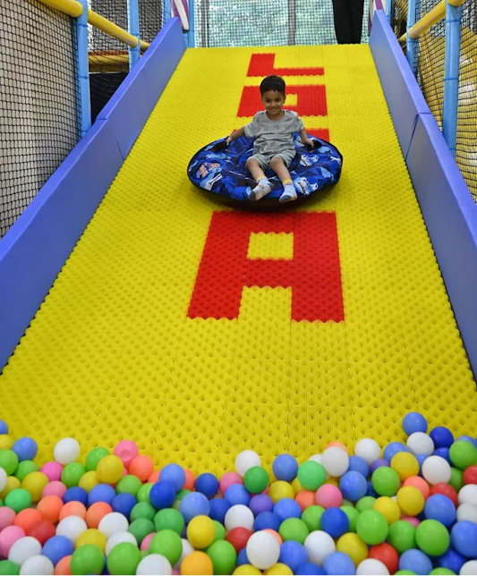 A young boy slides down a yellow and red mat-covered slide on a blue inflatable, landing in a ball pit.