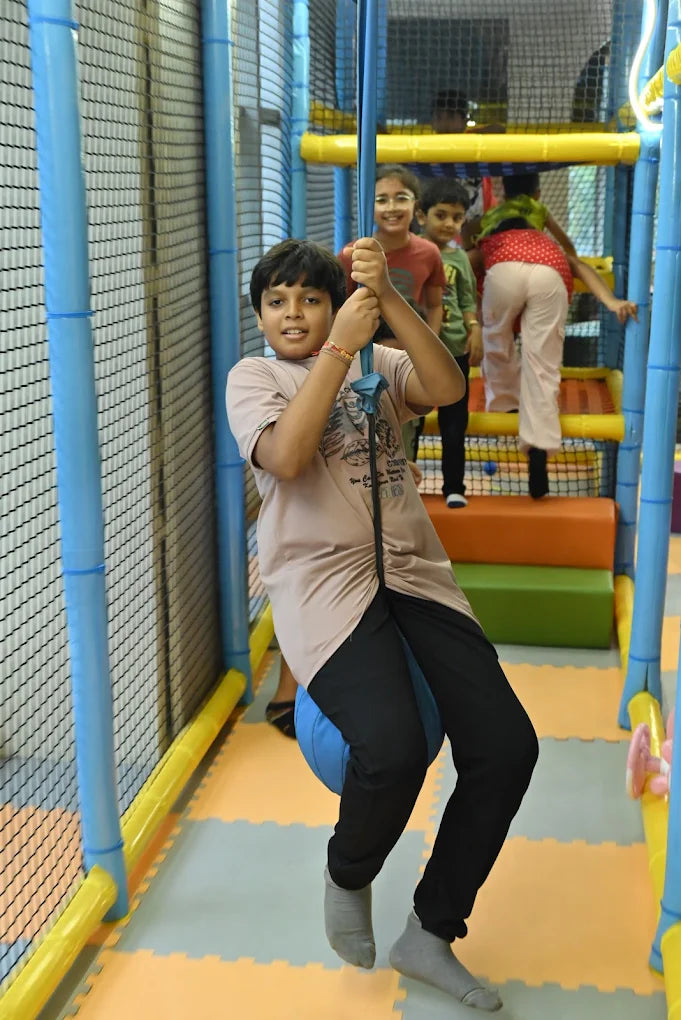 A young boy swings on a blue ball in a colorful indoor playground, with other children in the background.